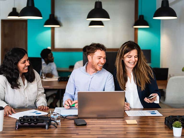 Three professionals sitting at a desk with an open laptop working at a shared co-working space and smiling as woman holds up phone to share information with the other two people.